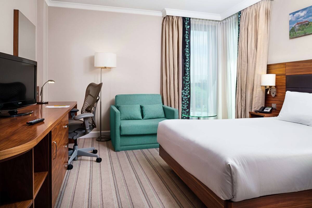 A hotel room with a bed, a desk with a TV, a green armchair, a lamp, and a window with curtains.