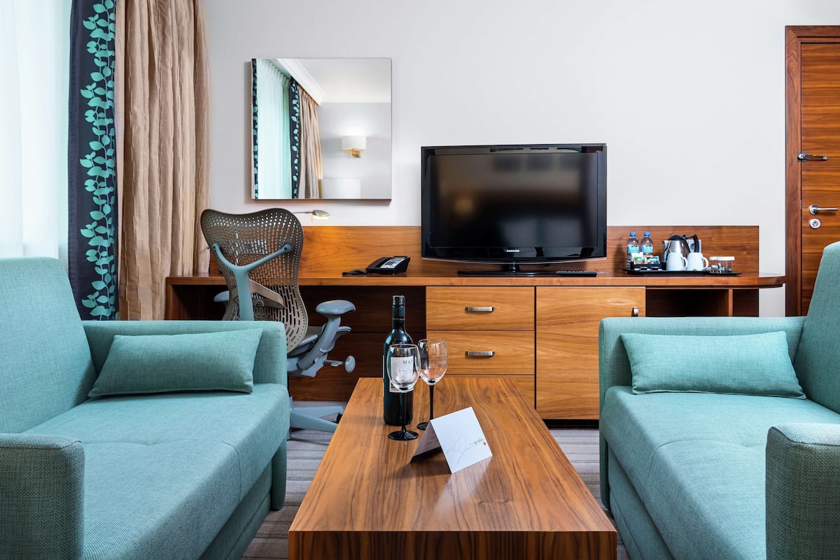 A hotel room with a wooden desk, two teal sofas, a television, and a minibar.