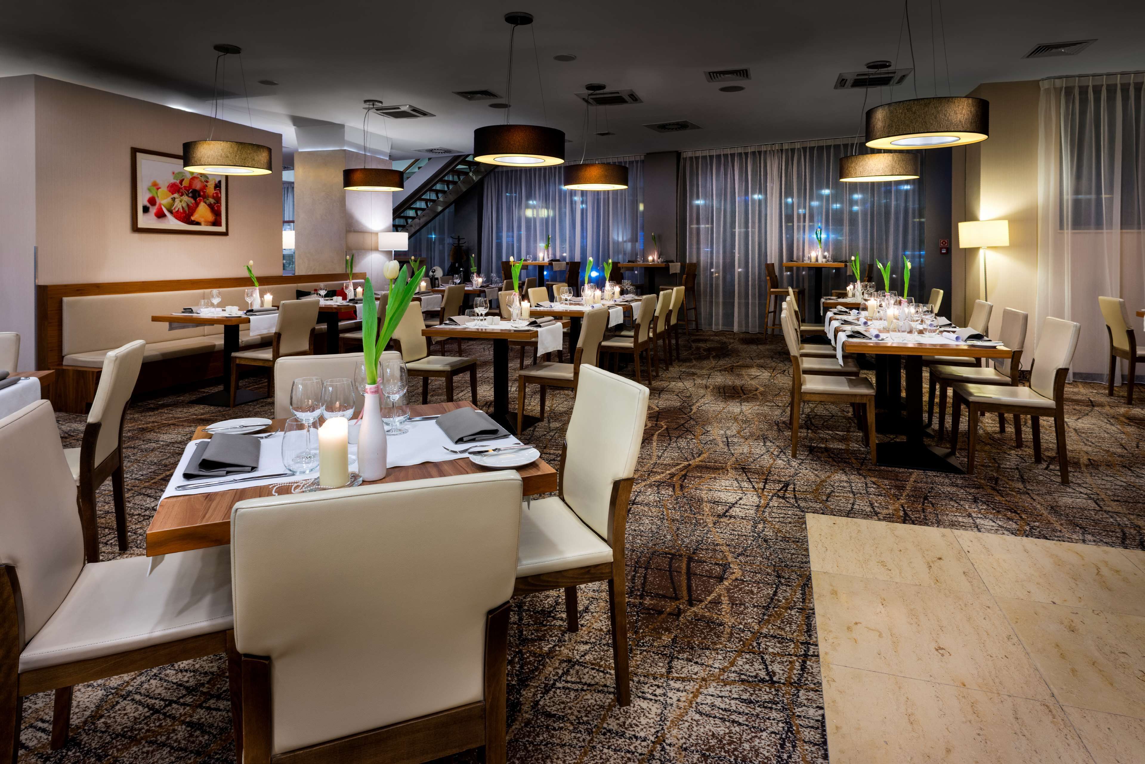 Een restaurantgedeelte van een hotel met meerdere tafels gedekt voor gasten, met moderne verlichtingsarmaturen en een vloerbedekking.