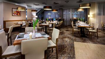 Een restaurantgedeelte van een hotel met meerdere tafels gedekt voor gasten, met moderne verlichtingsarmaturen en een vloerbedekking.