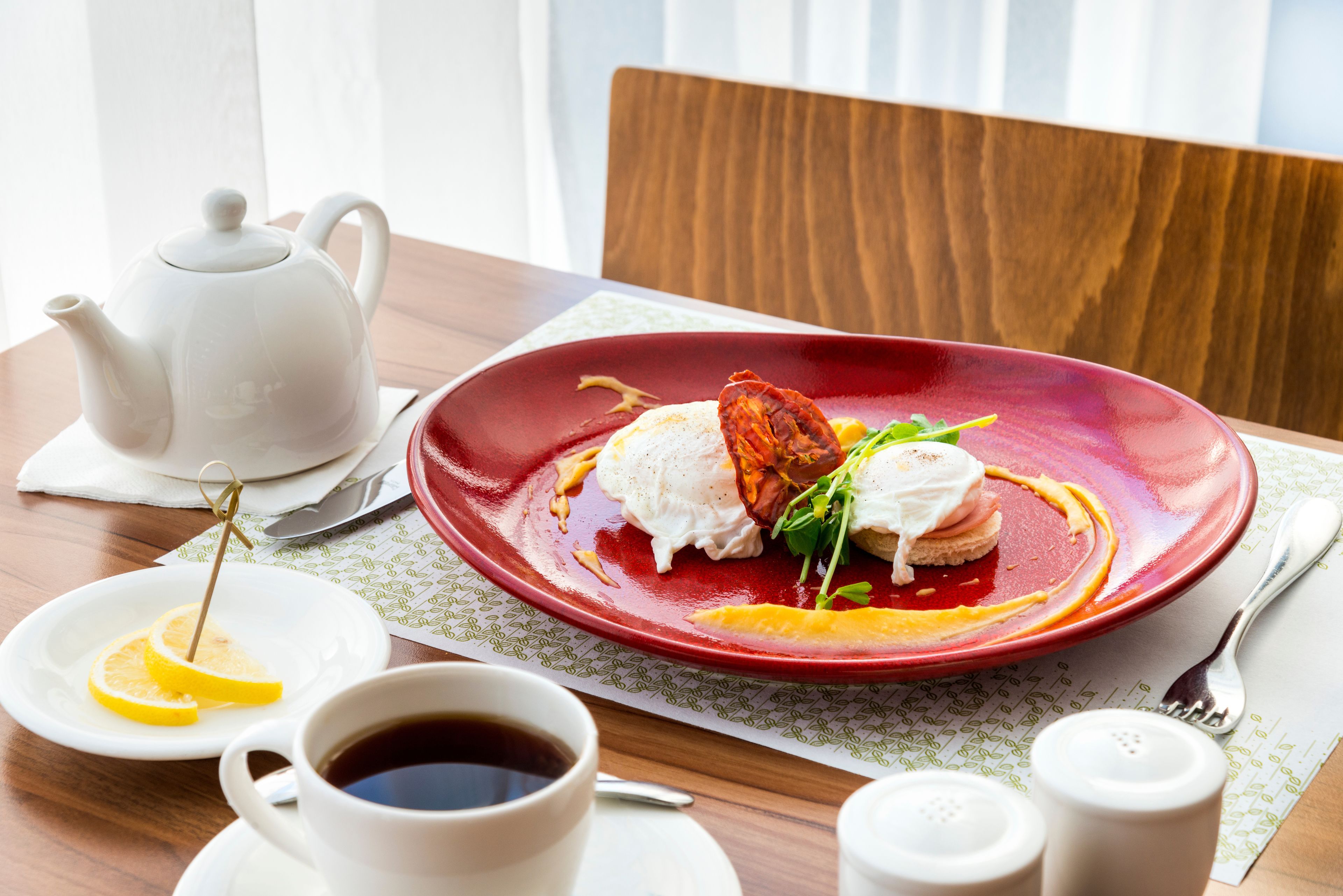 A dining table with a red plate of food, a white teapot, a cup of coffee, and salt and pepper shakers.