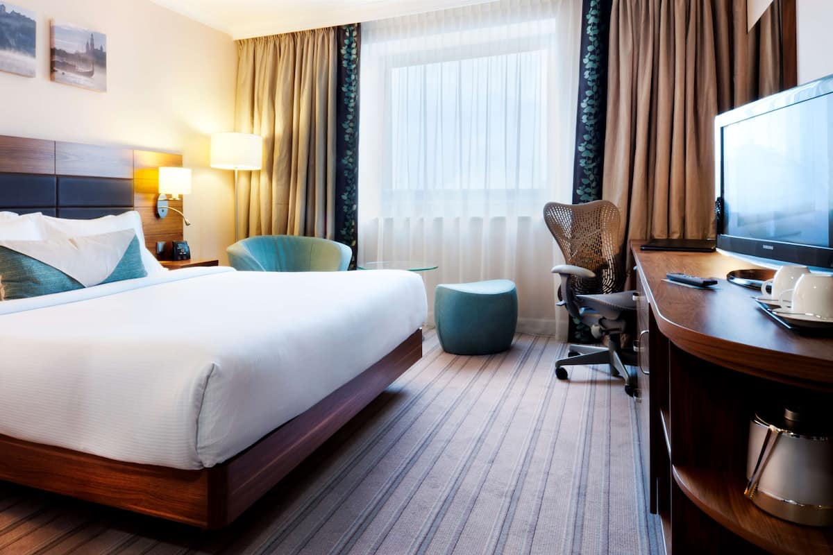 A hotel room with a large bed, a desk with a television, a chair, and a small ottoman.