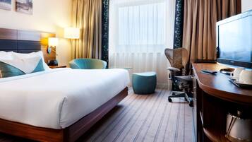 A hotel room with a large bed, a desk with a television, a chair, and a small ottoman.