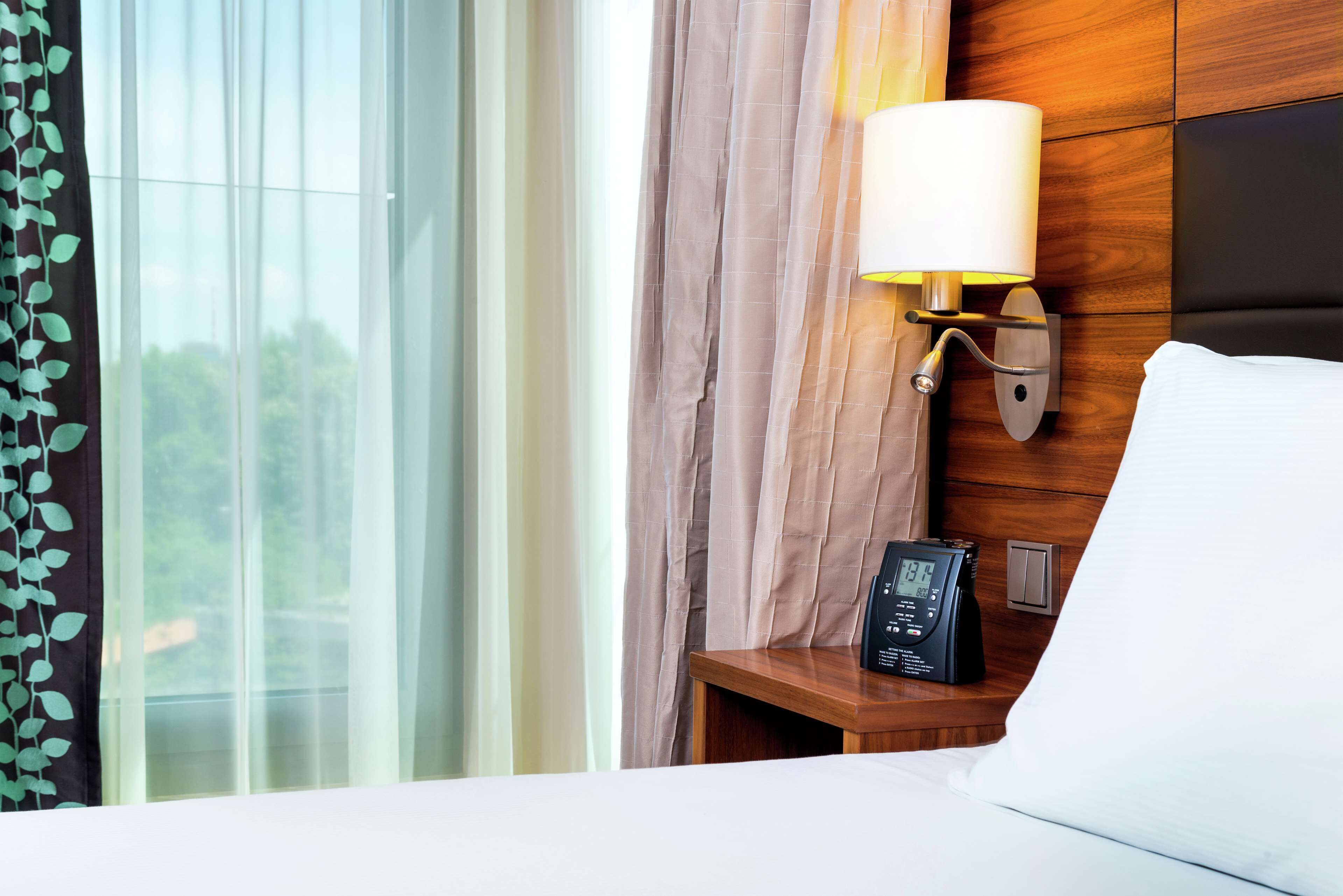 Una habitación de hotel con cama, una mesita de noche con lámpara, un reloj digital y una ventana con cortinas.