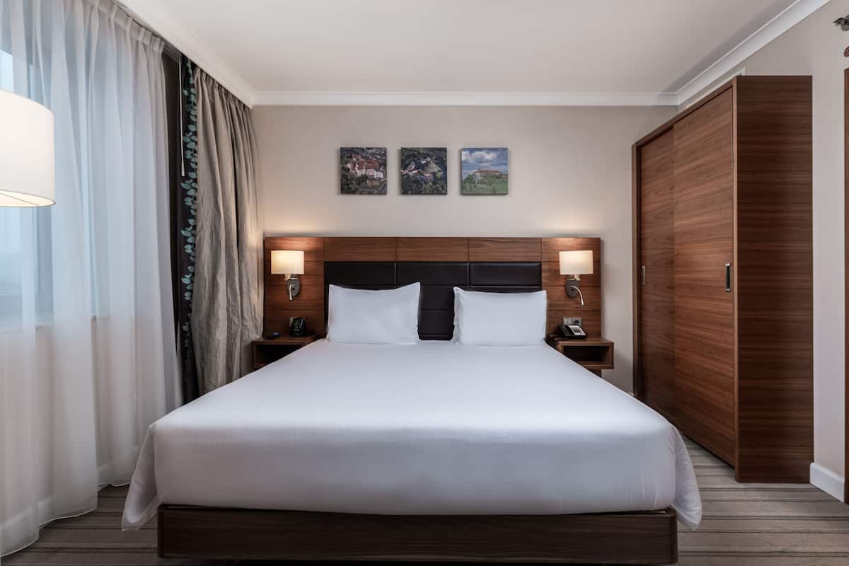 A hotel room with a large bed, wooden headboard, two bedside tables with lamps, a wardrobe, and three framed pictures on the wall.