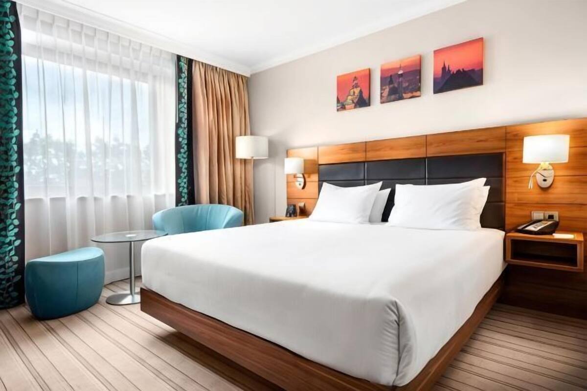 A hotel room with a large bed, two bedside lamps, a small table, a blue armchair, and a view of the city through the window.