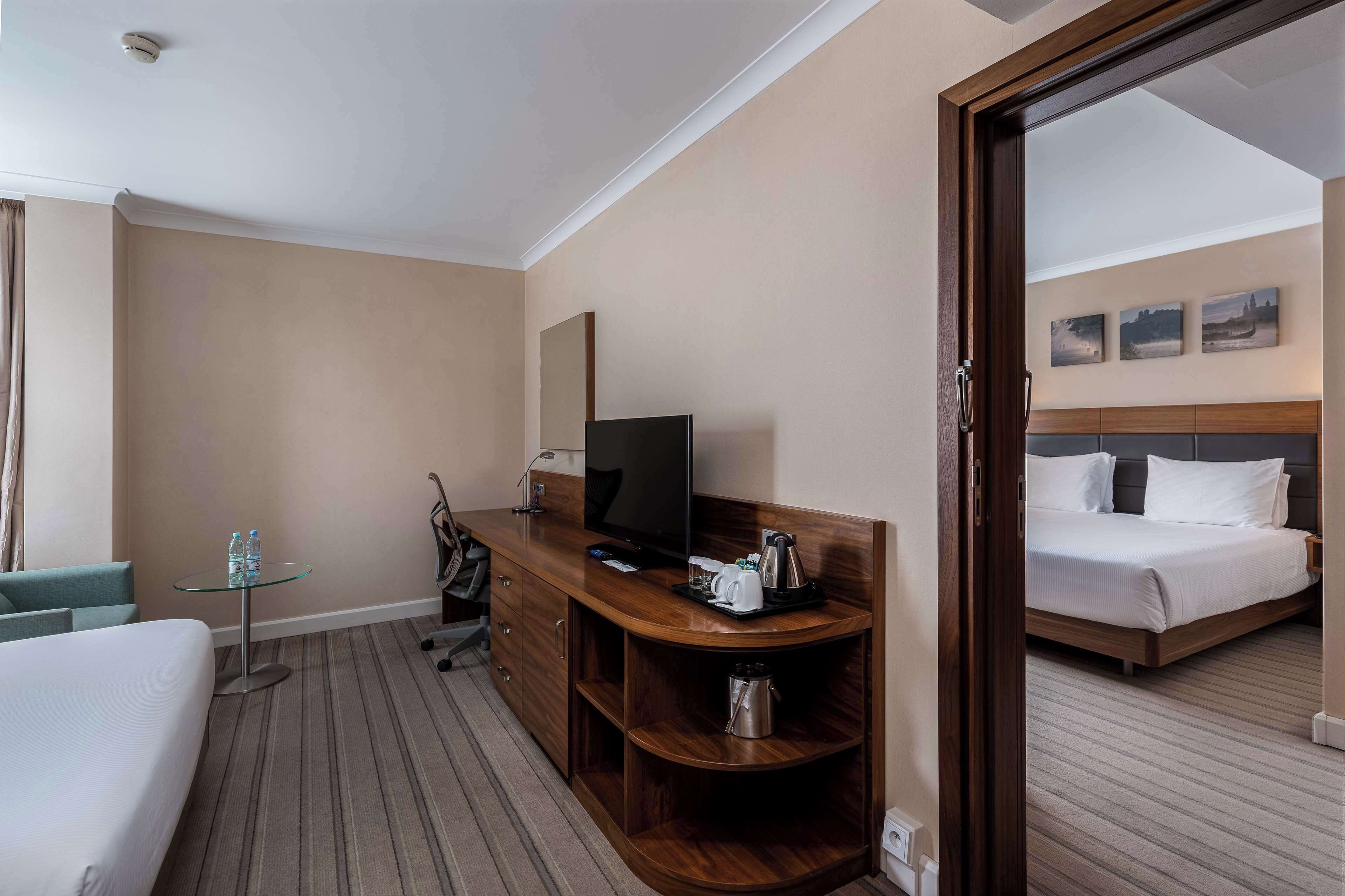 Habitación de hotel con cama, televisión, escritorio con cafetera y vista a otra habitación.