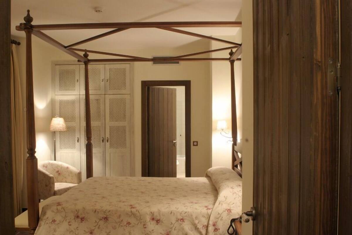 Ein Bett mit Baldachin, eine blumige Tagesdecke, ein Holzstuhl und ein Kleiderschrank mit Klappen.