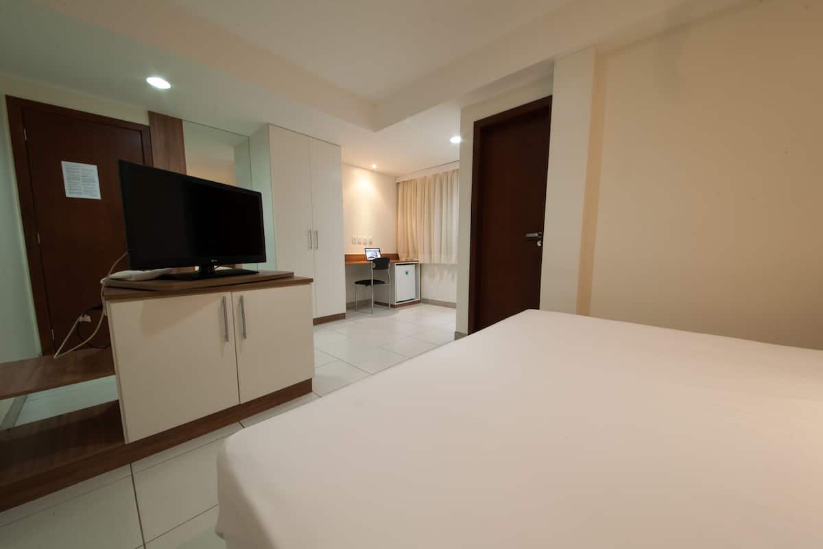 A hotel room with a bed, a TV on a wooden cabinet, a desk with a chair, and a bathroom visible through an open door.