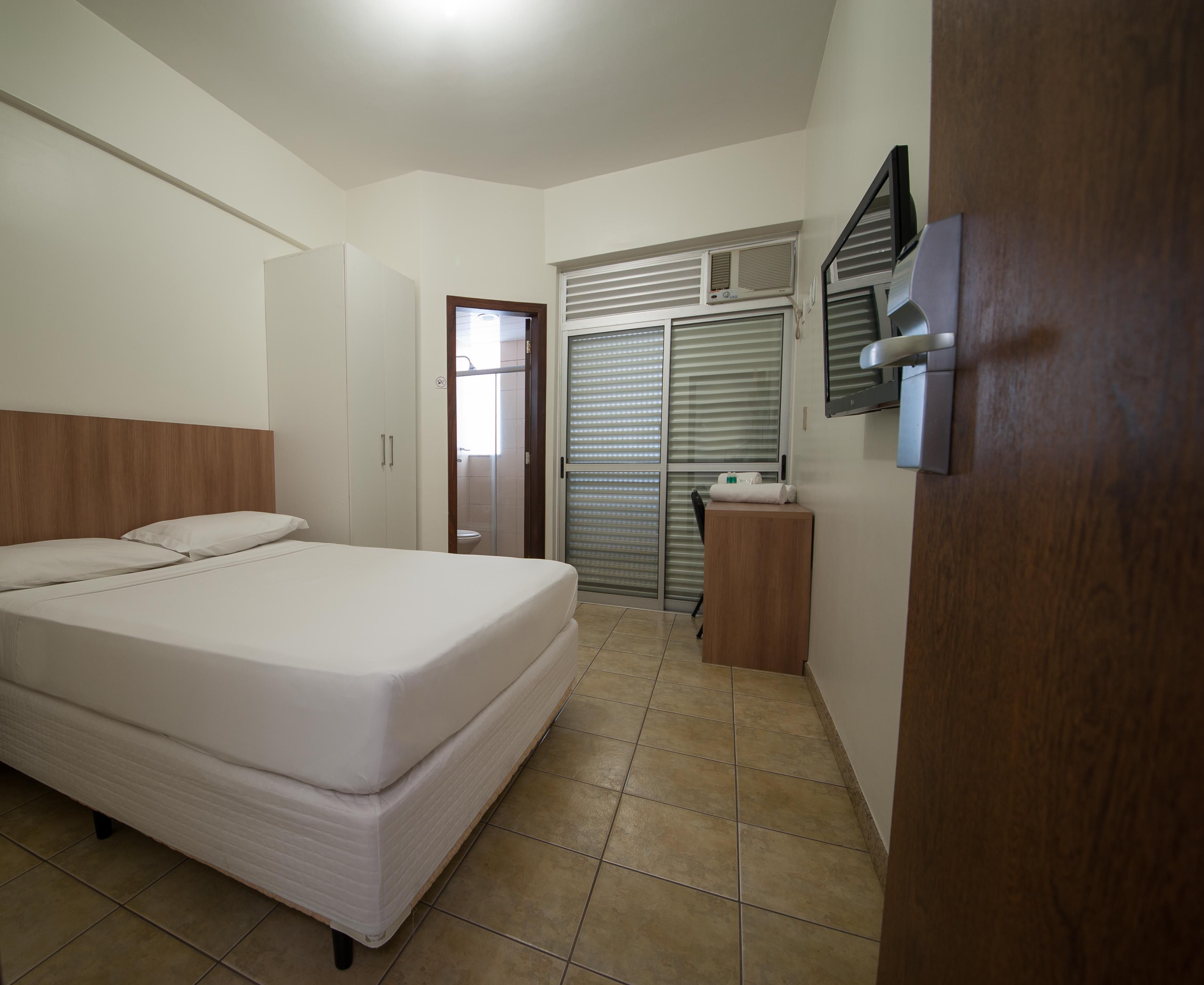 A bedroom with a bed, a wooden headboard, a wardrobe, a bathroom with a toilet, and a window with blinds.