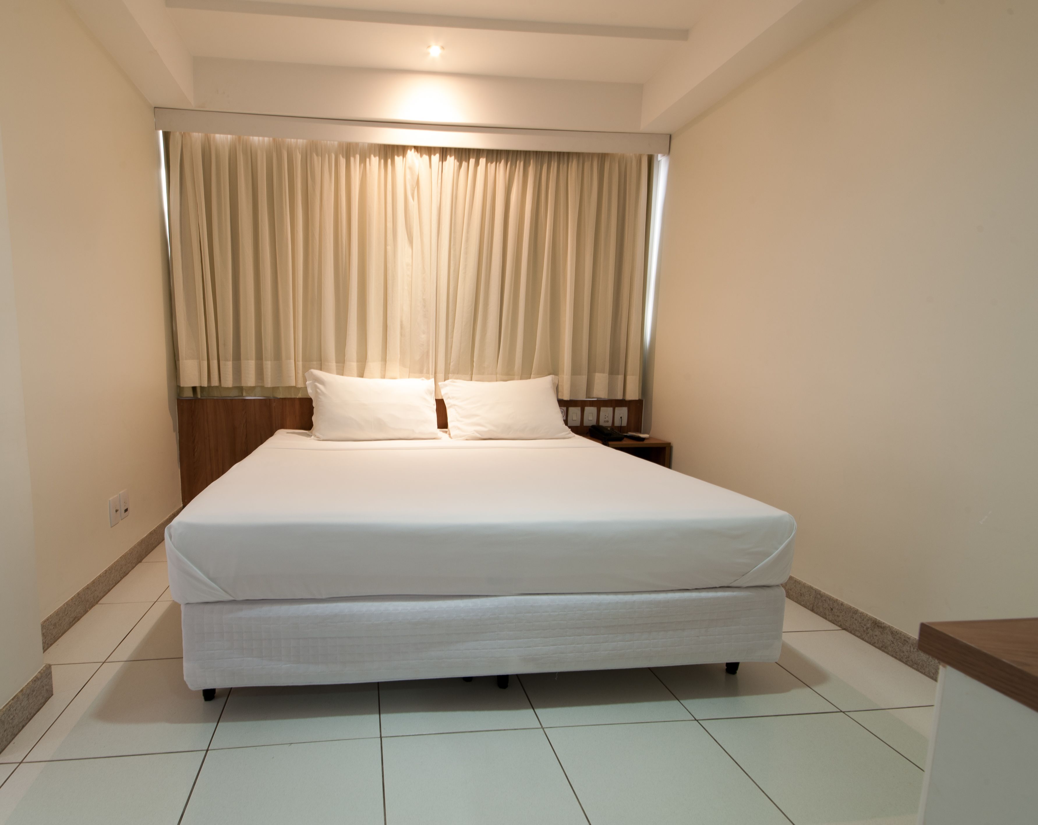 Una cama bien tendida con cabecera, una mesita de noche con lámpara, y una ventana con cortinas.