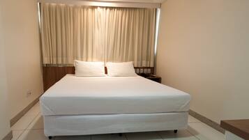 Una cama bien tendida con cabecera, una mesita de noche con lámpara, y una ventana con cortinas.