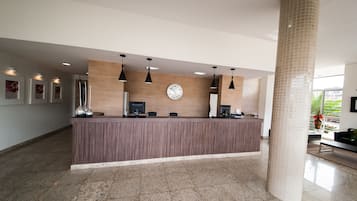 Uma área de recepção com balcão de madeira, vários monitores de computador, iluminação pendente, um relógio e um pilar grande.
