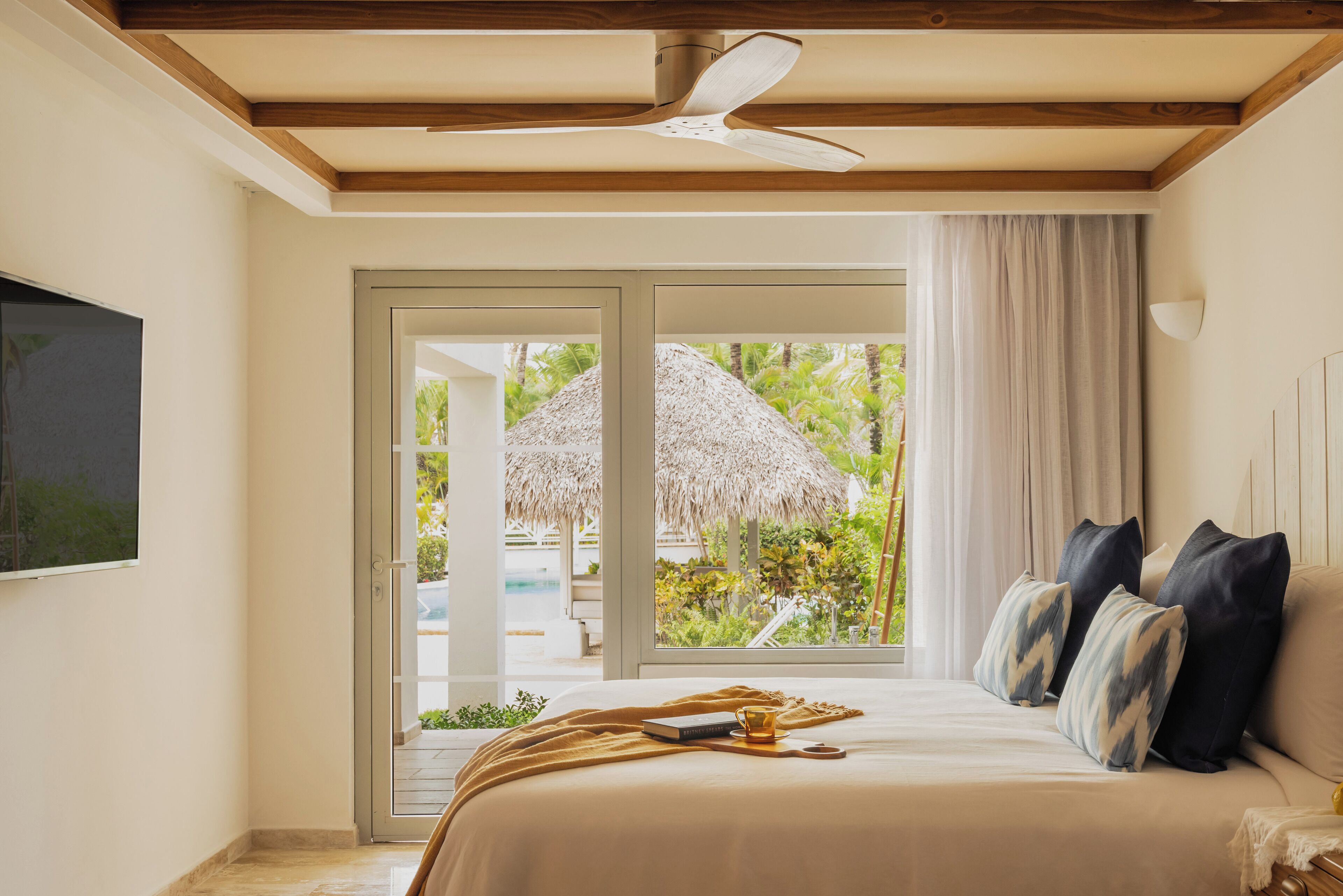 Un dormitorio con cama, ventilador de techo, televisor y vistas a una piscina y una construcción con techo de paja.