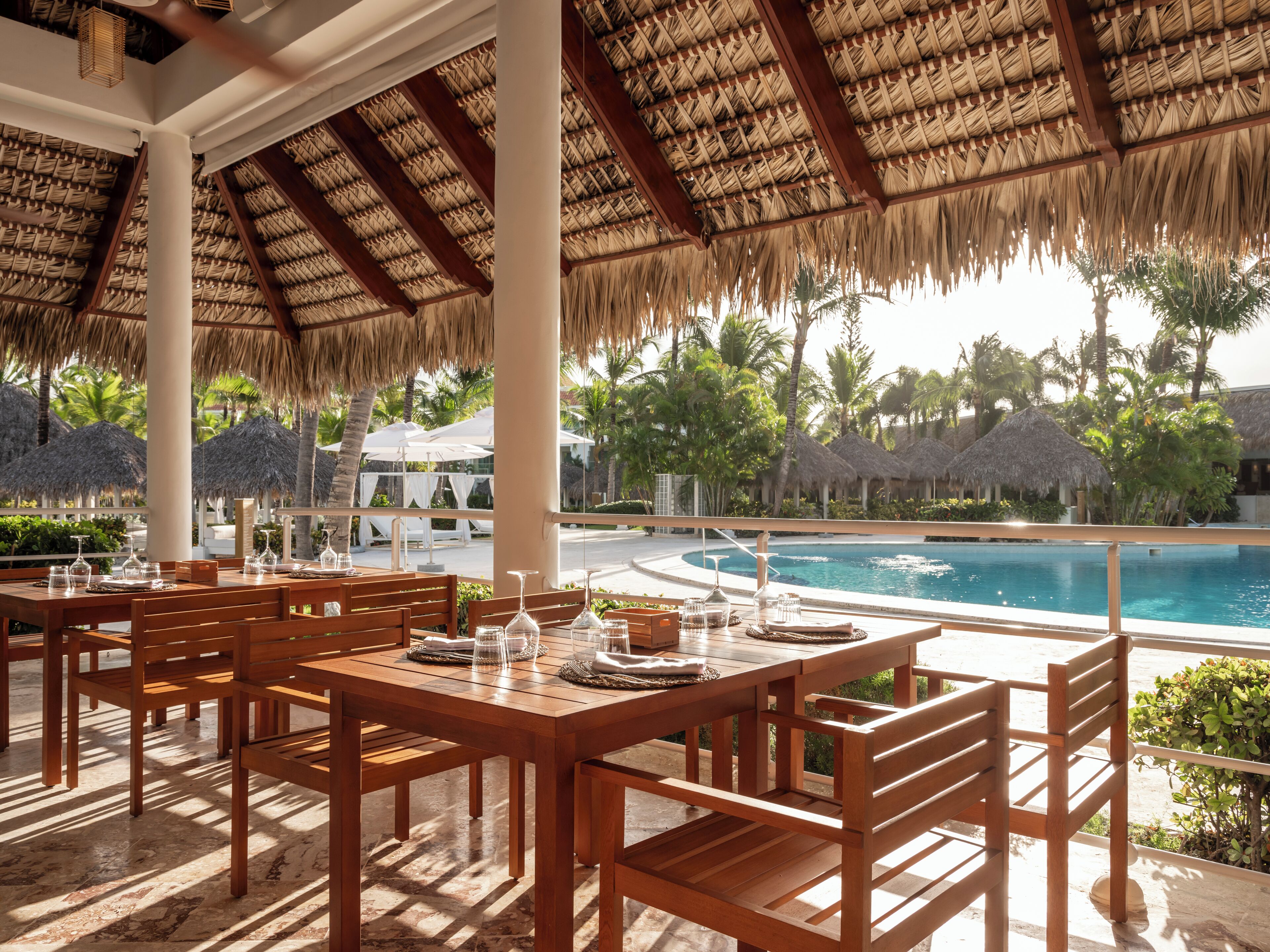 A poolside dining area with wooden tables and chairs, a thatched roof, and a view of a swimming pool and palm trees.