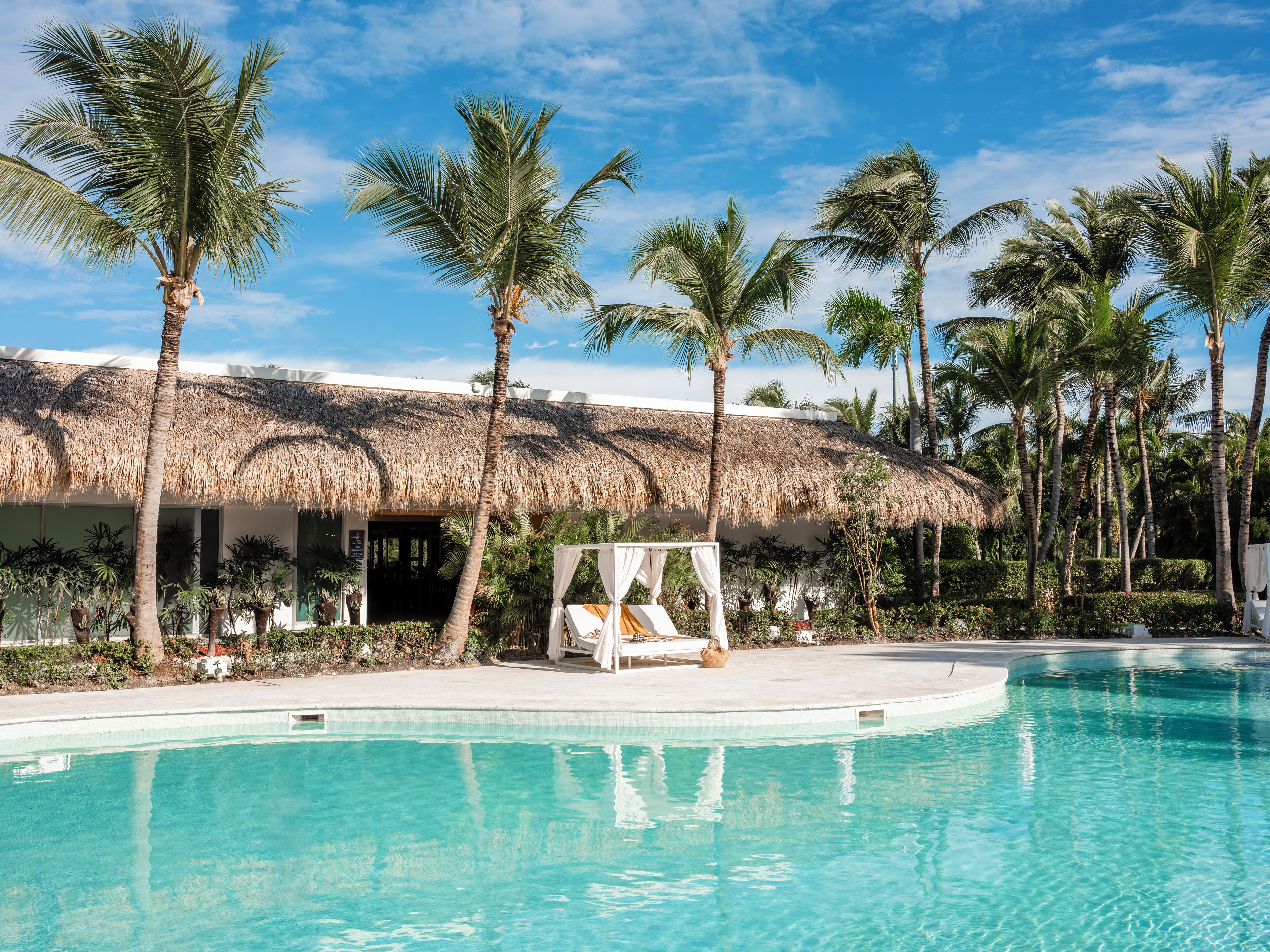 Zona de piscina con un edificio de techo de paja, palmeras y una silla de descanso.
