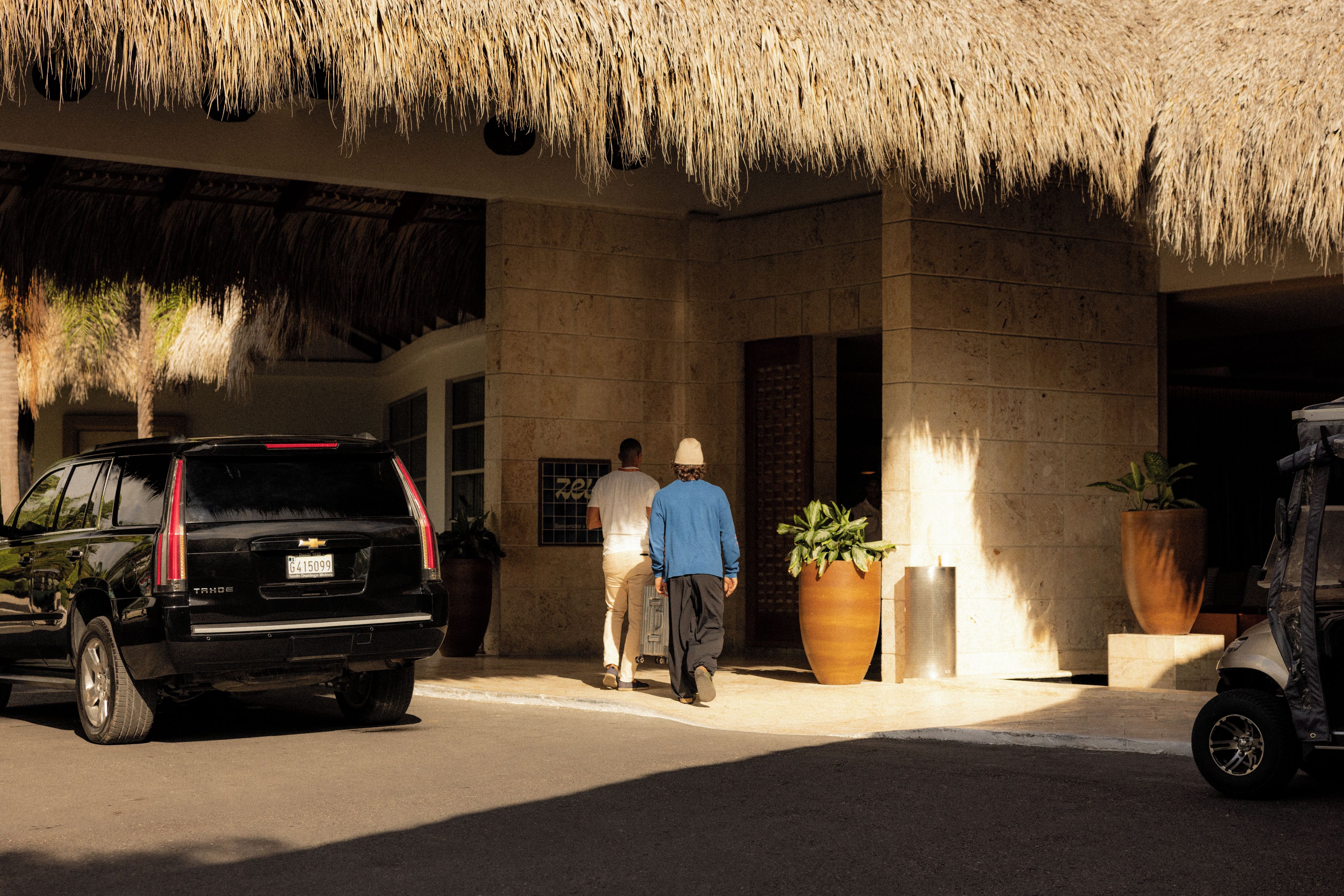 Una entrada de edificio con techo de paja, un SUV estacionado y grandes macetas con plantas.