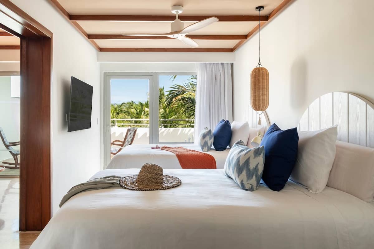 Habitación de hotel con cama, ventilador de techo, televisor, balcón con vista a palmeras y una lámpara de mimbre.