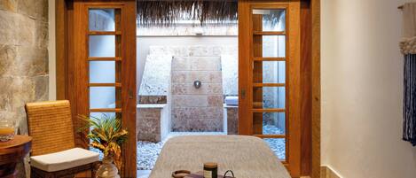 A spa room with a massage table, a wooden chair, a small table with a drink, and a view of a stone wall and a thatched roof structure.