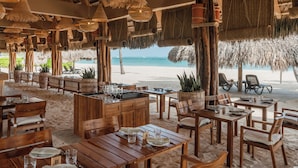 En restaurant ved stranden med træborde og -stole, stråparasoller og udsigt over havet.