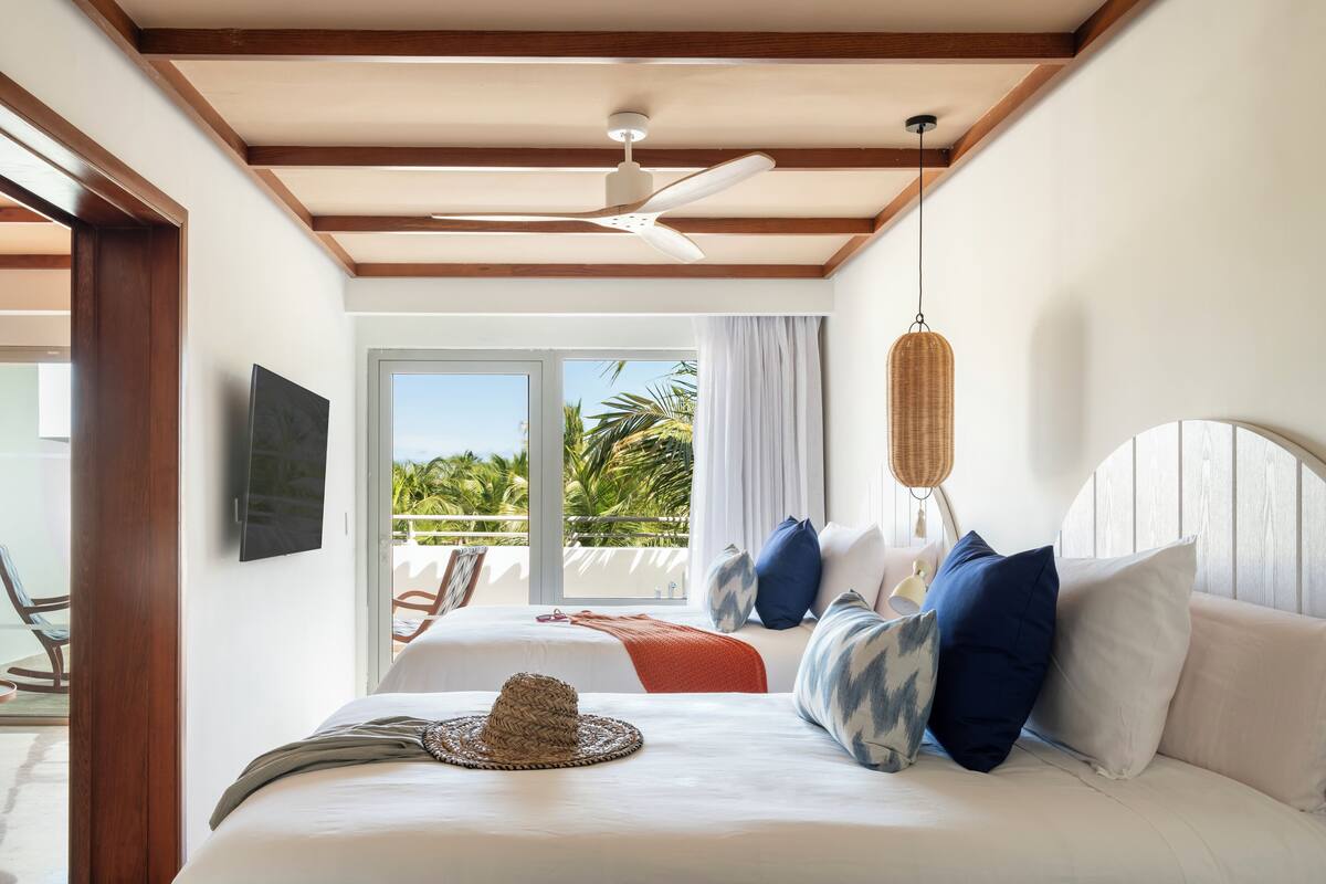 Habitación de hotel con cama, ventilador de techo, televisión y balcón con vista a palmeras.