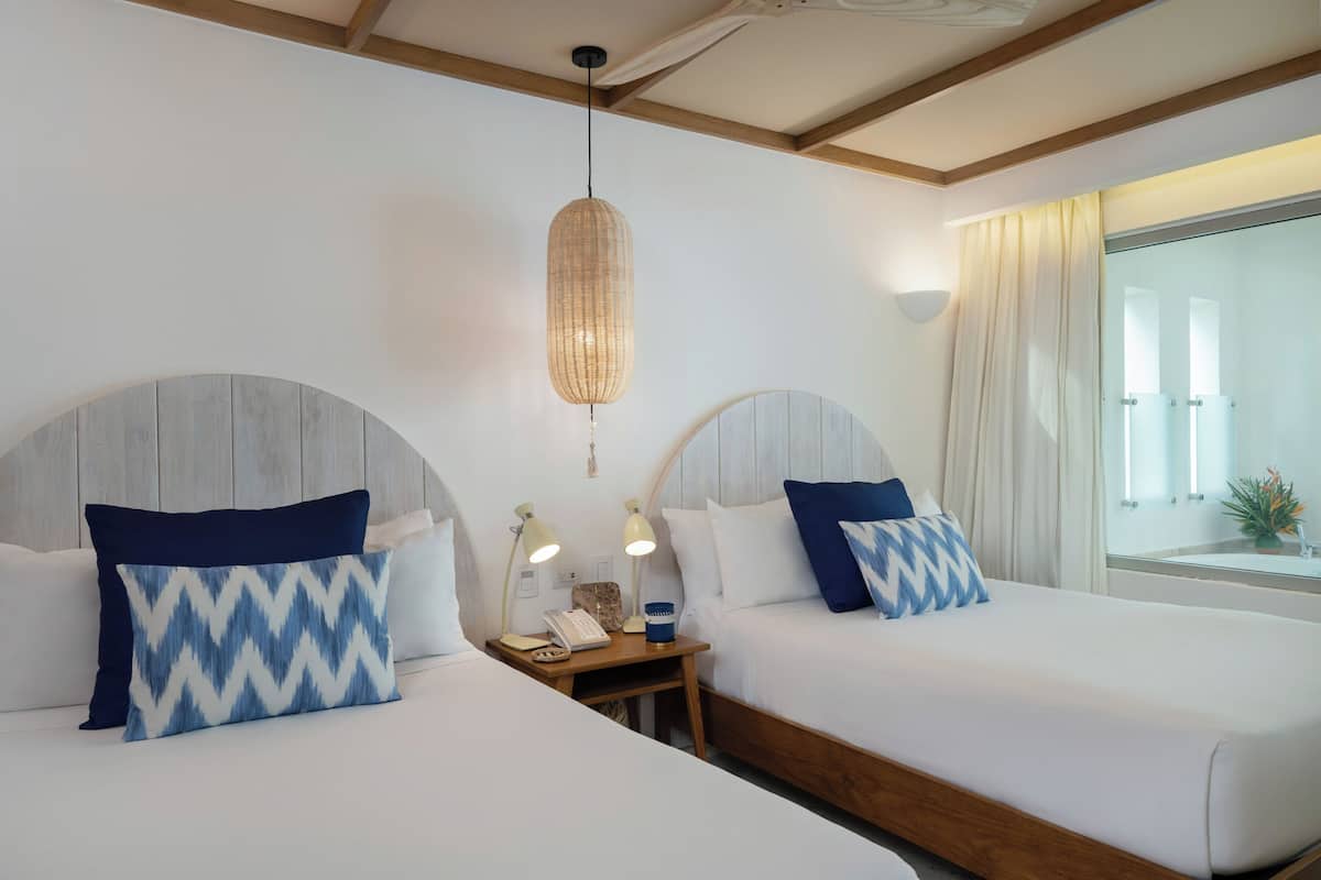 Habitación de hotel con dos camas, cada una con cabecera, mesita de noche con lámpara y ventana con cortinas.