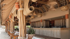 A beachside bar with wooden tables and benches, a thatched roof, and a variety of hanging lights. - Zel Punta Cana, All Suites - All inclusive (Punta Cana)