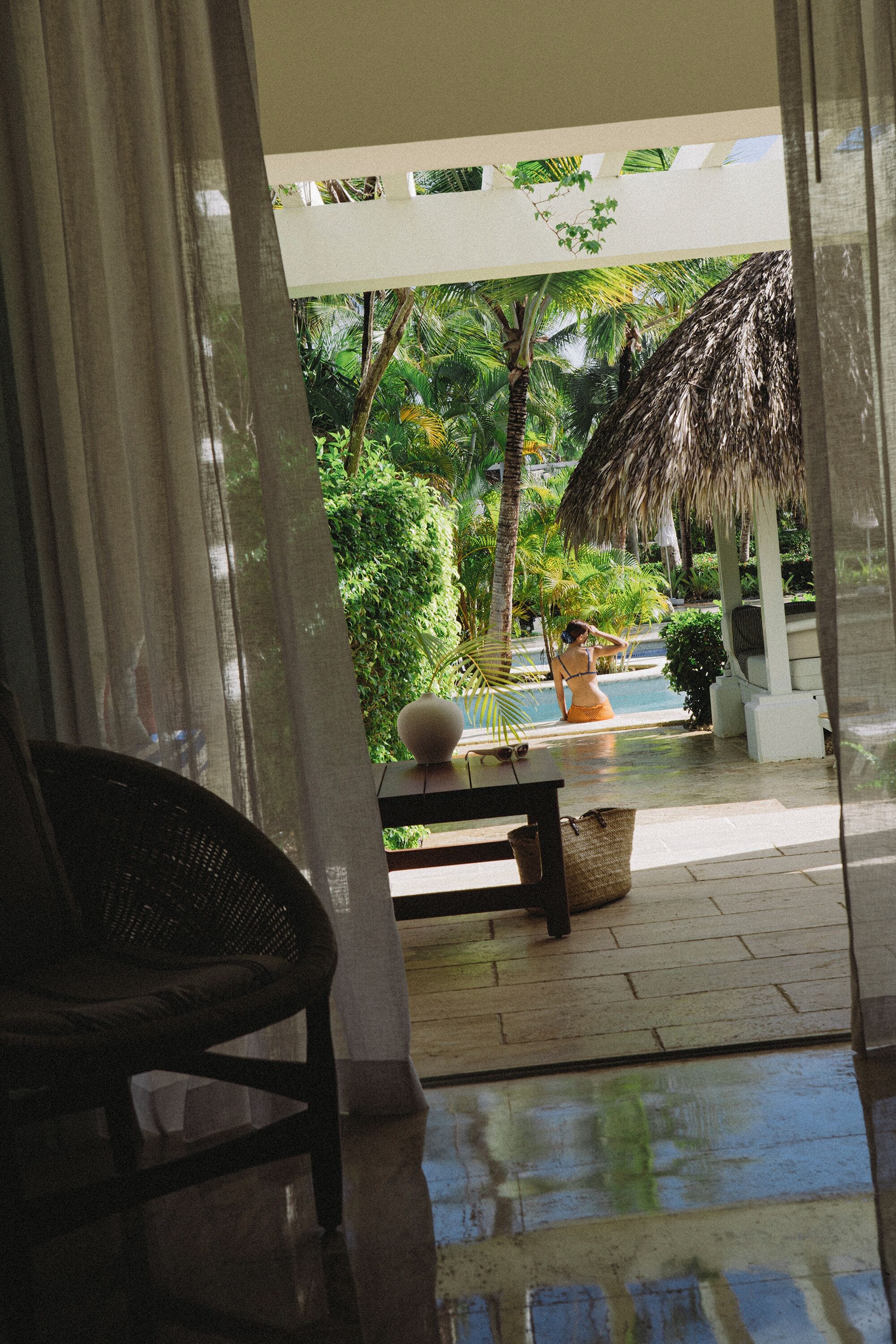 A view through a window with a chair, a table, and a pool area with a thatched roof and greenery.