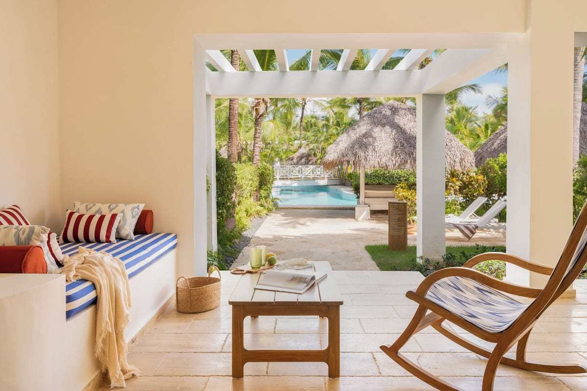 Una habitación con vista a una piscina, una mecedora de madera, un sofá con cojines a rayas y una pequeña mesa con un libro y un vaso.