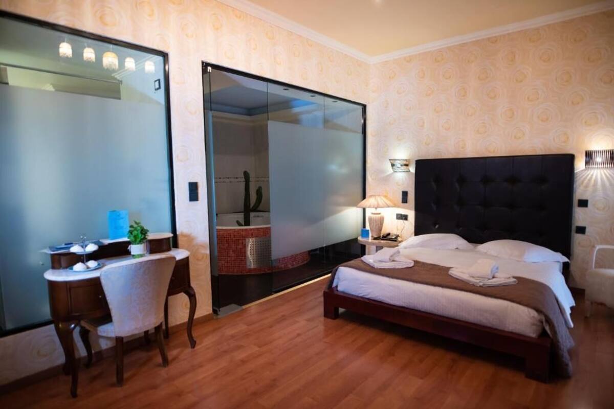 Habitación de hotel con cama, escritorio, silla, espejo y baño visible a través de una puerta de cristal.