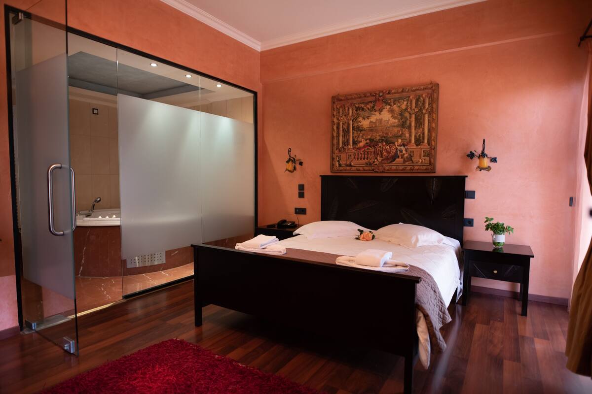 Un dormitorio moderno con cabina de ducha de vidrio, una cama grande con ropa de cama blanca, un cuadro clásico en la pared y una mesita de noche con una planta.
