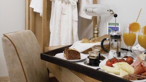 Une chambre d’hôtel avec une chaise en bois, un peignoir suspendu à un porte-manteau, un plateau de petit-déjeuner avec du pain, du fromage, des tomates et une cafetière.