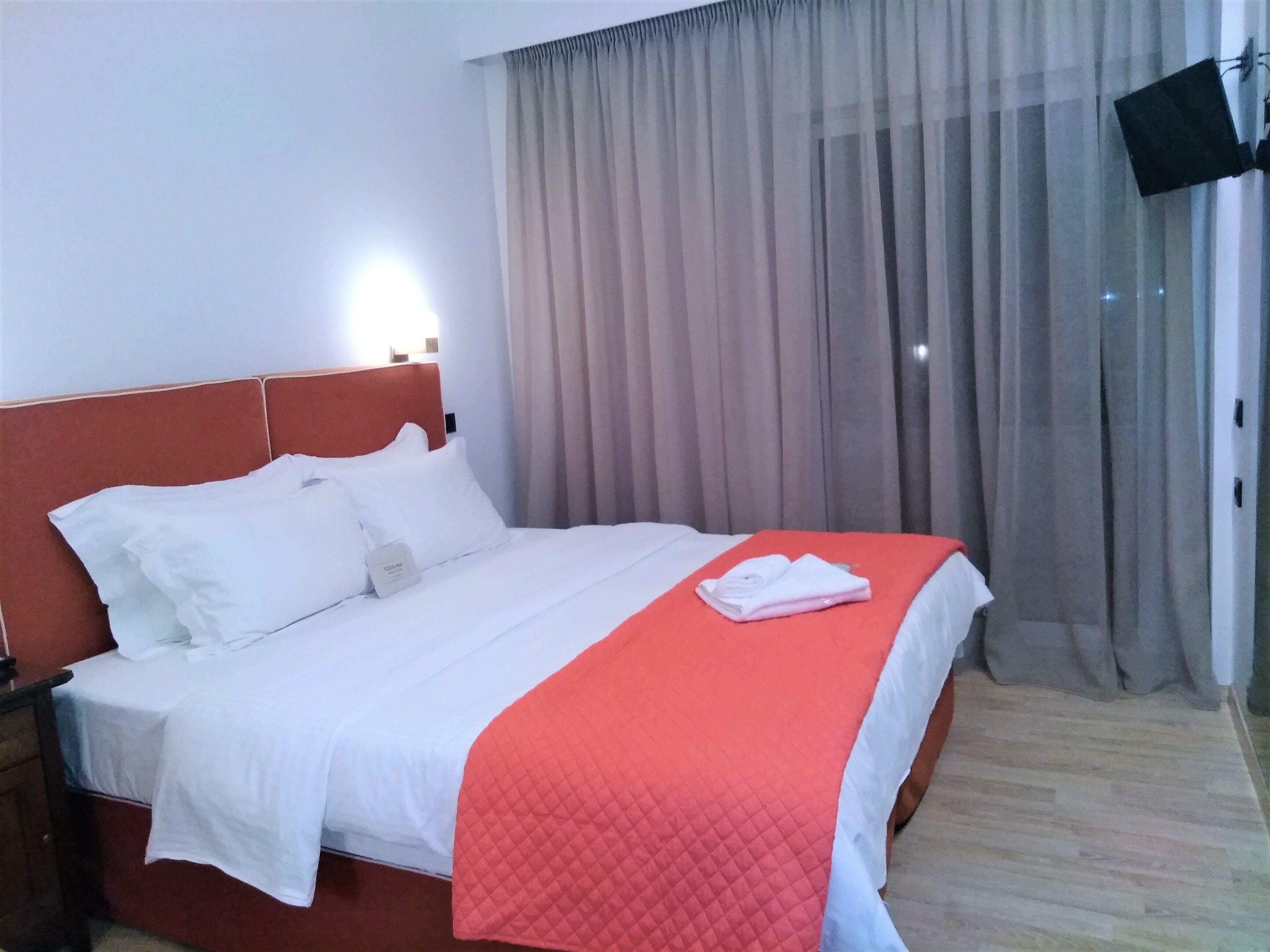 A neatly made bed with white linens and a red quilted blanket, a wooden nightstand with a lamp, and a large window with sheer curtains.