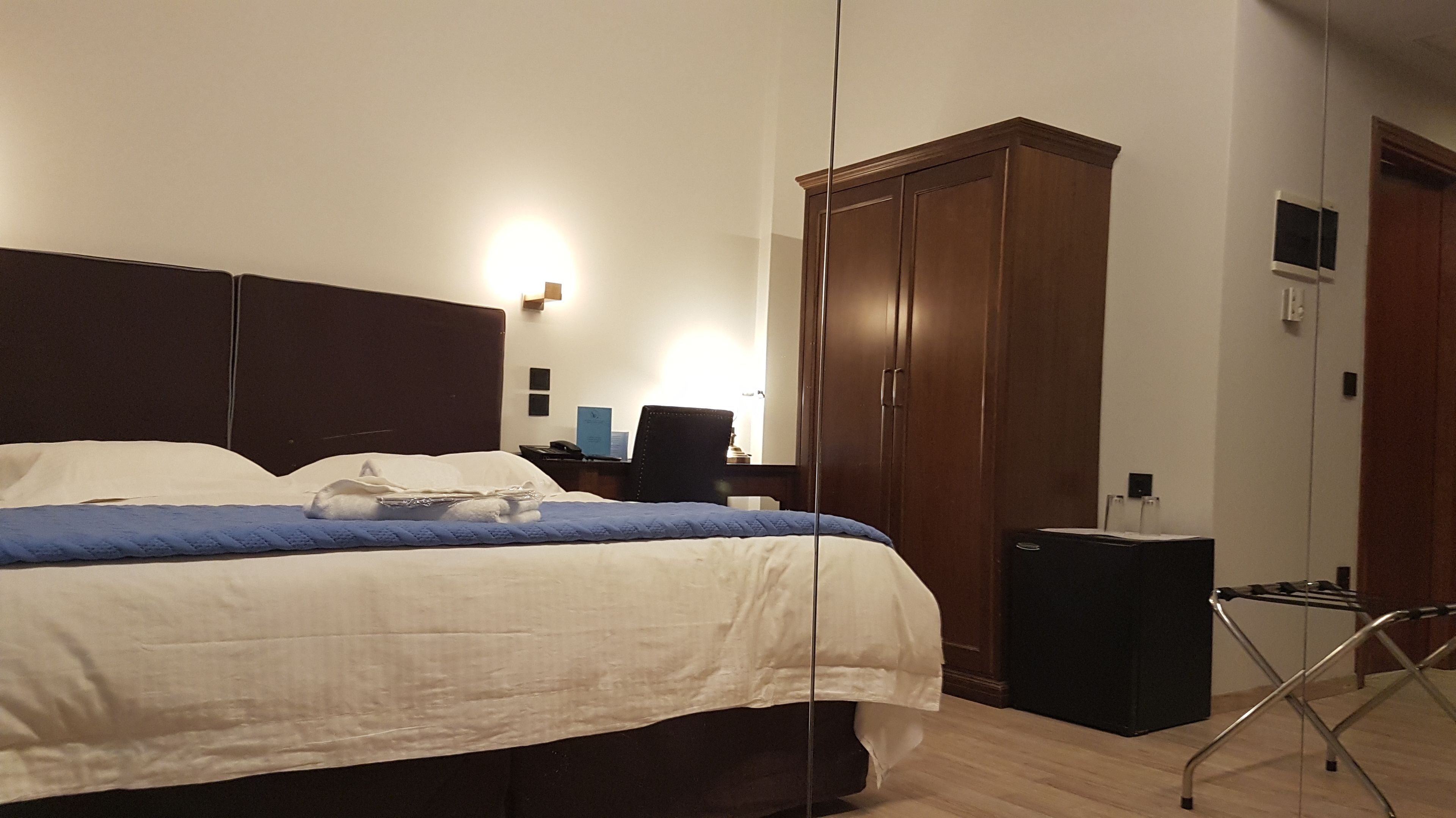 Une chambre d’hôtel avec un grand lit, une armoire, un bureau avec une chaise et une petite table avec une lampe.