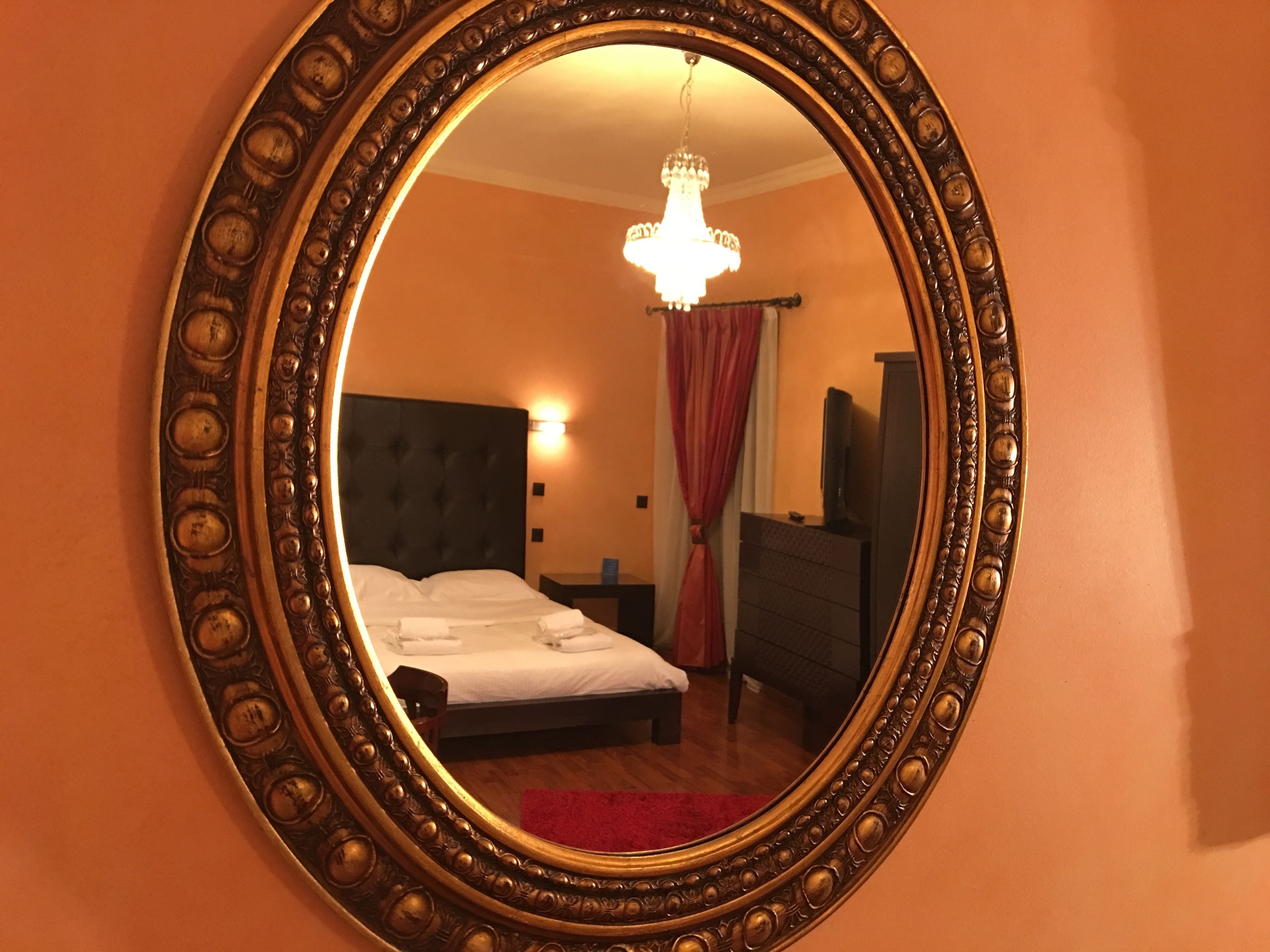 Quarto com um espelho grande e ornamentado, cama com roupa de cama branca, tapete vermelho, lustre e uma televisão sobre uma cômoda.