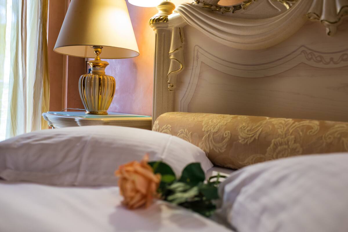 Una cama impecable con sábanas blancas, una sola rosa y una lámpara sobre una mesita de noche.