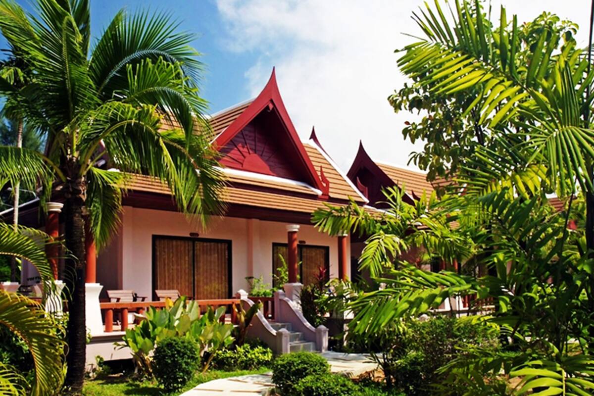 A traditional Thai villa with a red roof, surrounded by lush greenery and palm trees.