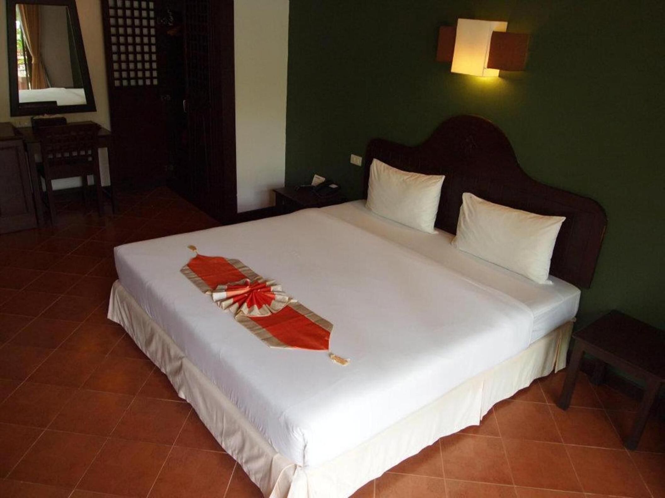 Una cama bien tendida con un cubreleche rojo y blanco decorativo, un cabecero de madera y una mesita de noche con una lámpara.