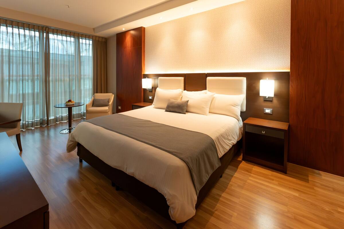 Ein Hotelzimmer mit einem großen Bett, Nachttischen, einem Sessel, einem kleinen Tisch und einem Fenster mit Vorhängen.