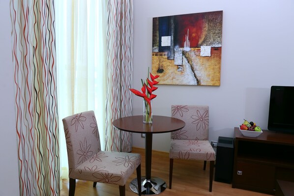 Una sala moderna con dos sillones de estampado floral, una mesa redonda con un jarrón de flores rojas, un cuadro en la pared y un frutero sobre un mueble de madera.