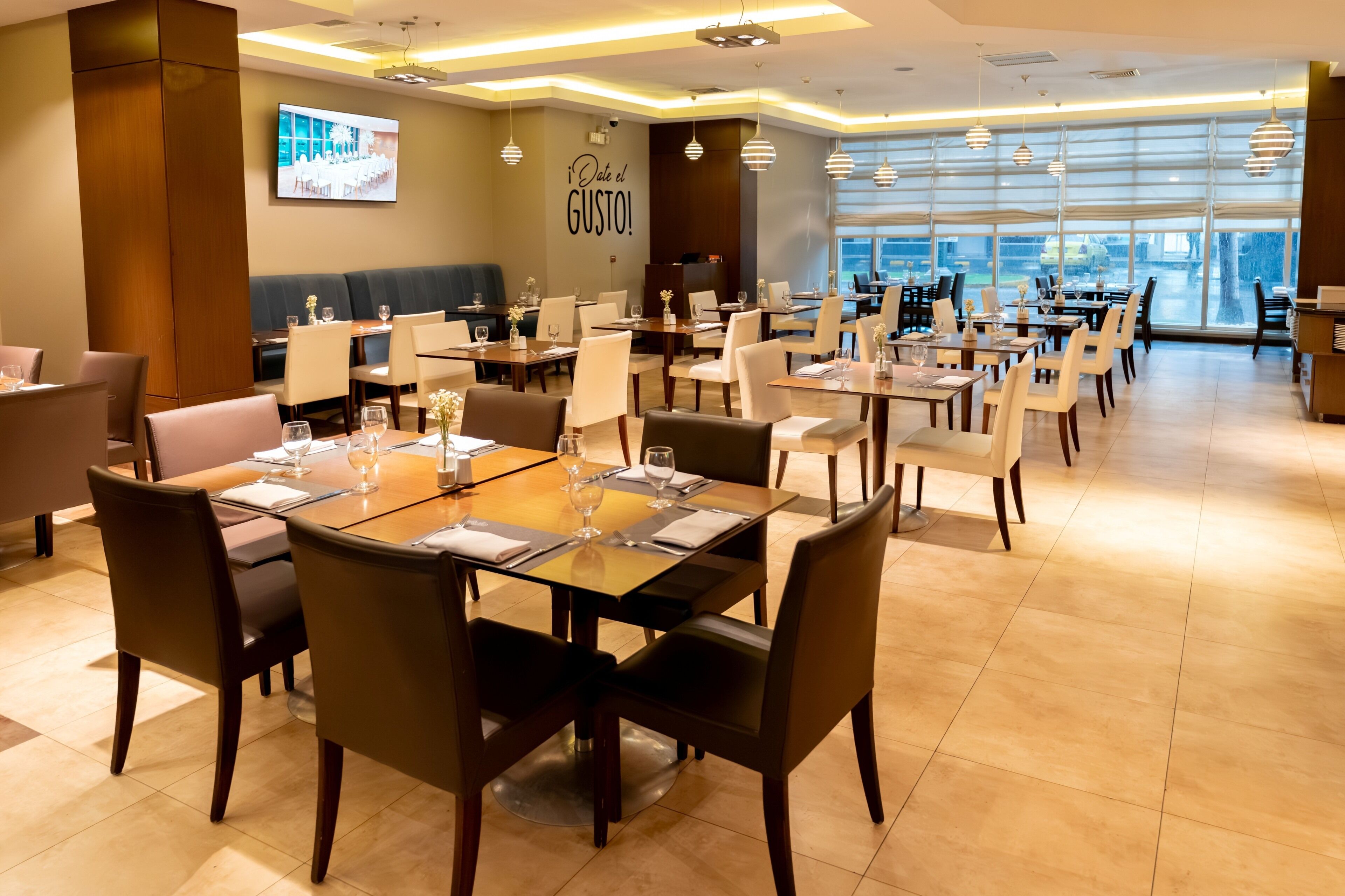 A modern dining area with multiple tables and chairs, a large screen displaying a wedding, and a wall with a motivational quote.