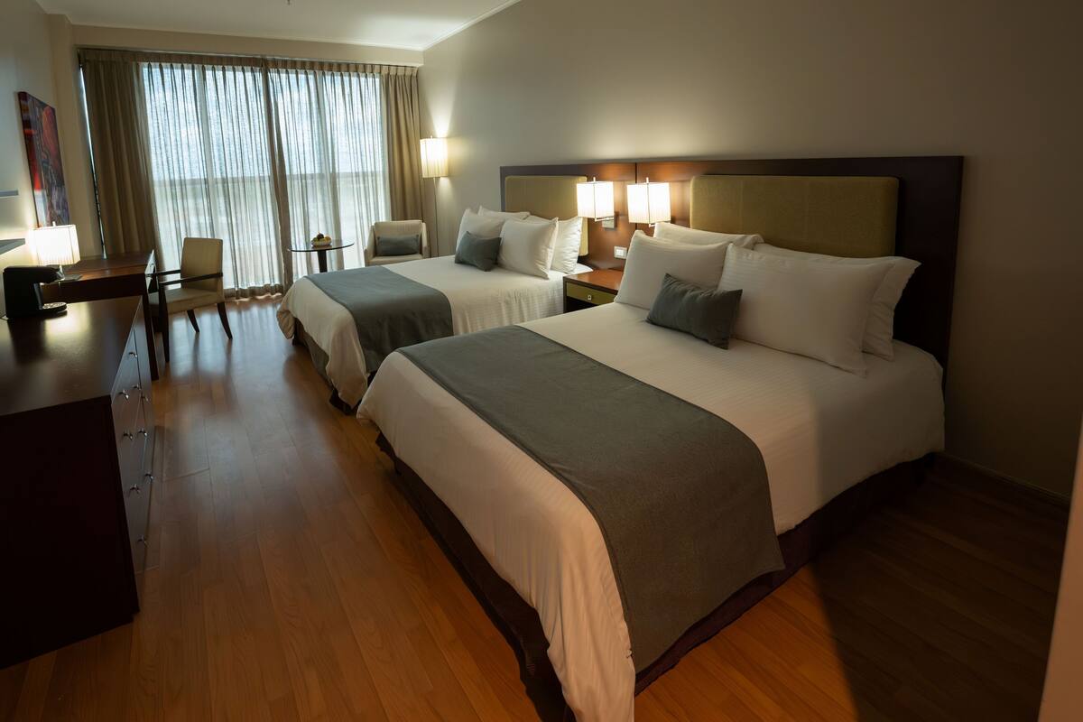 Ein Hotelzimmer mit zwei Betten, einem Schreibtisch, einem Stuhl, einer Lampe und einem fenster mit Vorhängen.