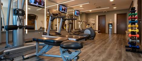 Une salle de sport équipée de machines de cardio, de matériel de musculation et d’un téléviseur diffusant une chaîne de sport.