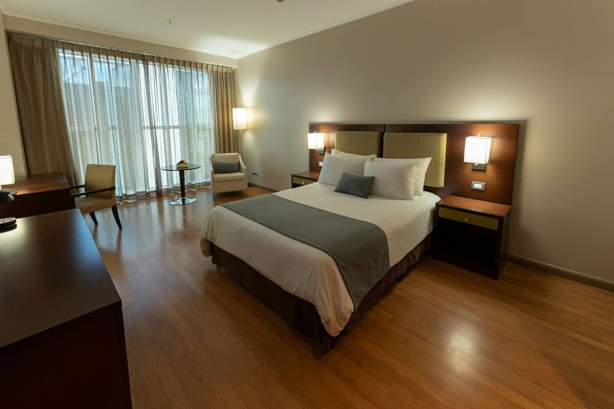 Ein Hotelzimmer mit einem großen Bett, Nachttischen, einem Schreibtisch, einem Sessel und Blick durch transparente Vorhänge.