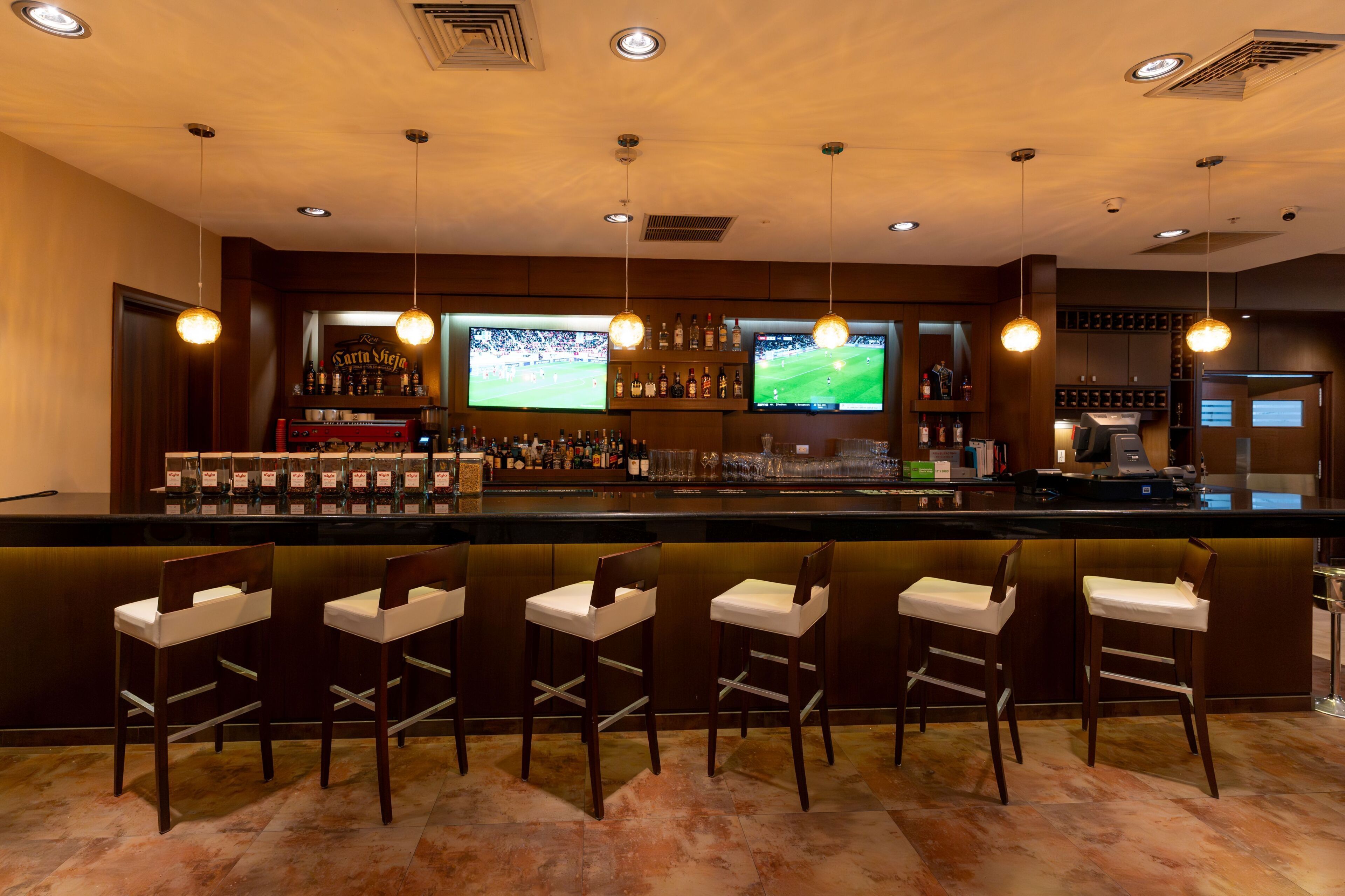 A bar area with a counter, multiple bar stools, a television screen, and overhead lighting.