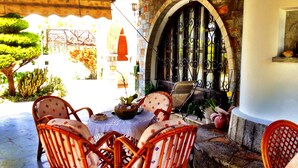 A covered patio with wicker chairs and a table, a stone wall with an arched window, and a cactus in the background. - Irene Villas (Ierapetra)