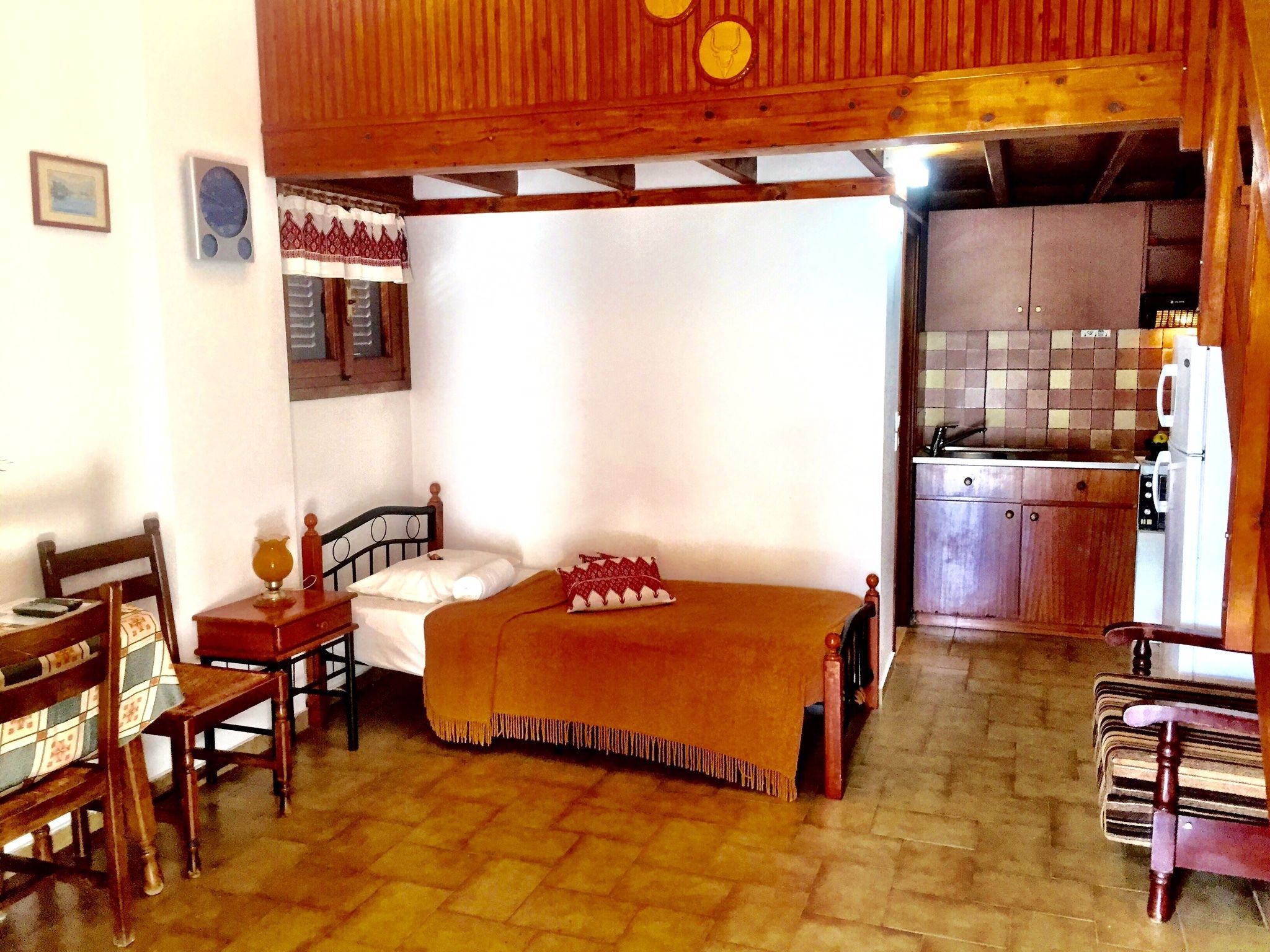 A room with a bed, a wooden chair, a small table, and a kitchen area with cabinets and a refrigerator.