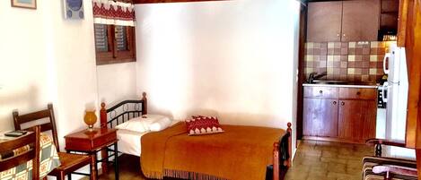 A room with a bed, a wooden chair, a small table, and a kitchen area with cabinets and a refrigerator.