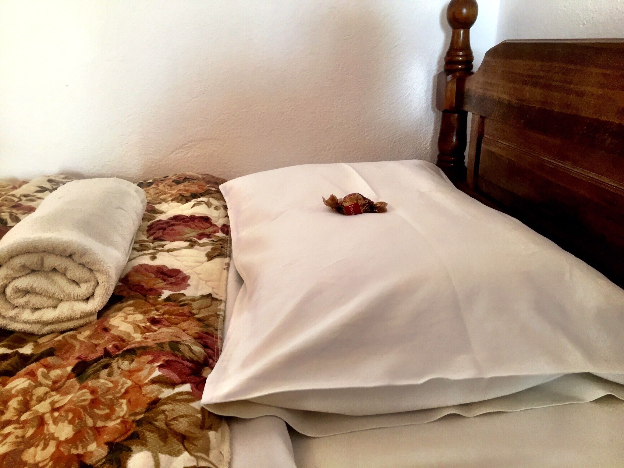 A bed with a floral-patterned bedspread, a neatly rolled white towel, and a small red object on the pillow.