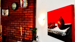 A brick wall with a clock and bookshelves, a table with a candle and a plant, and a red painting of fruits. - Irene Villas (Ierapetra)